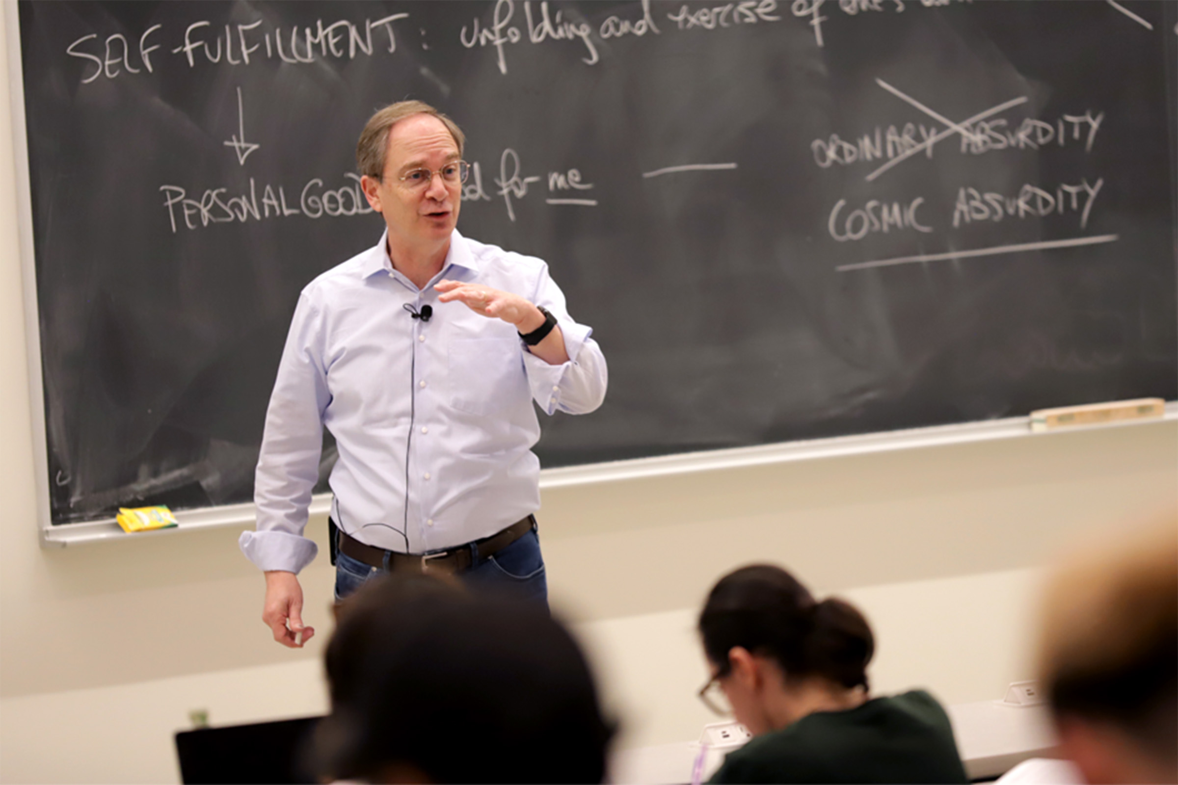 Person stands at front of classroom, speaking in front of a blackboard, as students look on.